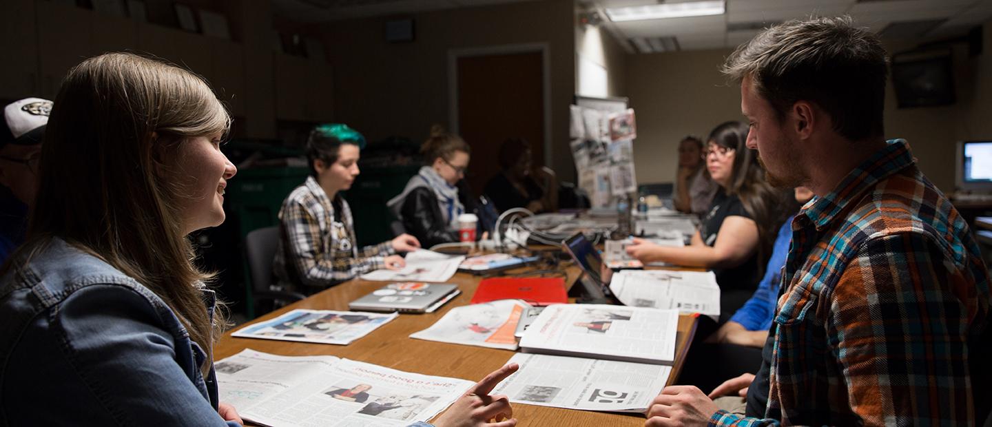 一群学生围着一张桌子坐着，桌子上放着打开的报纸